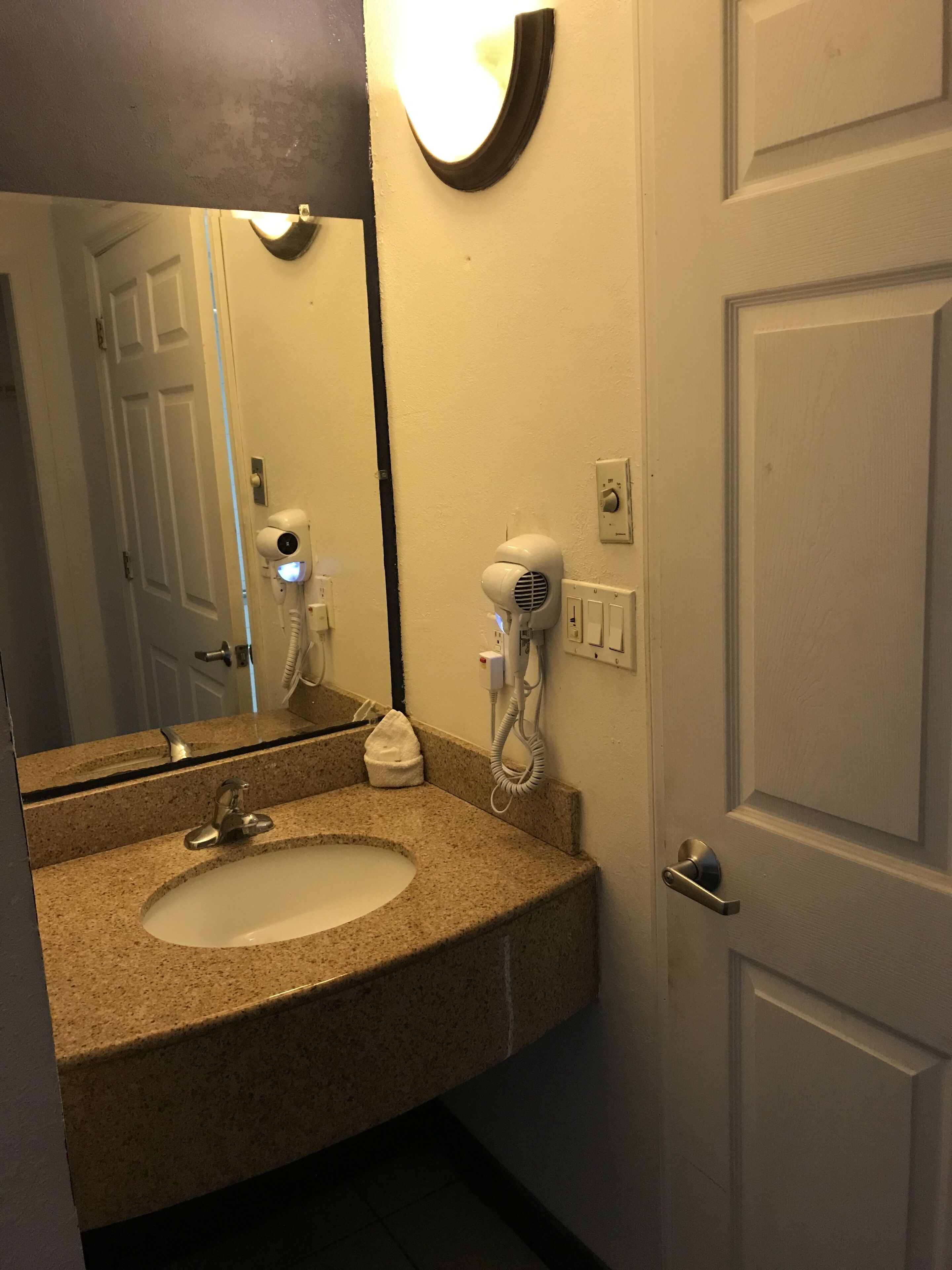 Un baño con lavamanos de encimera de granito, un espejo, un secador de pelo y una puerta.