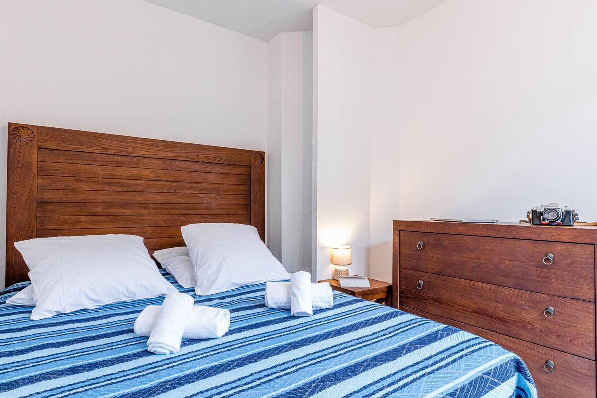 A neatly made bed with a striped blue and white bedspread, two white pillows, and two rolled towels on the bed.
