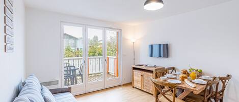 A bright living room with a sofa, a dining table with chairs, a TV mounted on the wall, and a sliding glass door leading to a balcony.