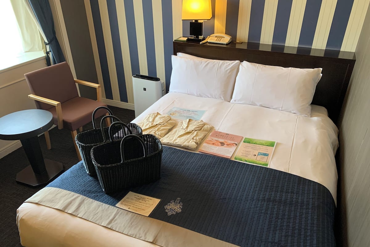 Ein Hotelzimmer mit einem ordentlich bezogenen Bett, einem Nachttisch mit Lampe, einem Sessel und einem Korb mit Pflegeprodukten.