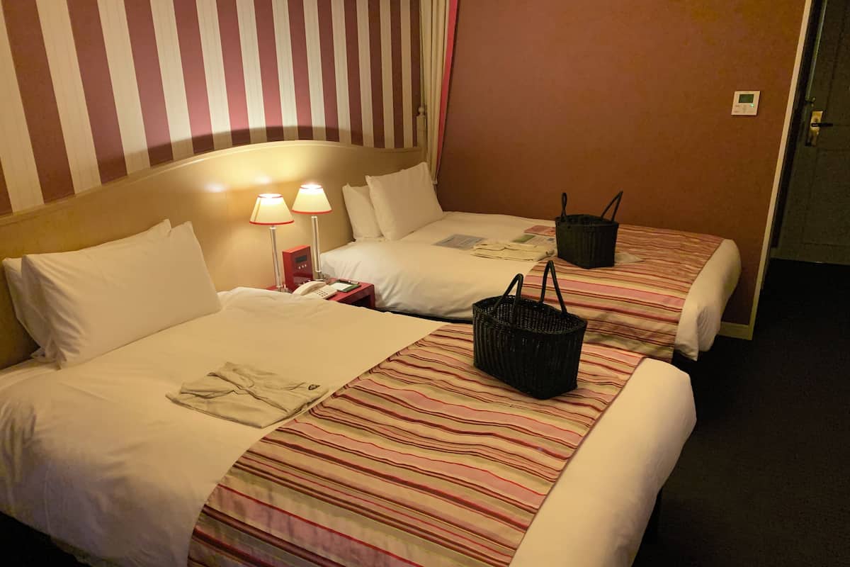 Ein Hotelzimmer mit zwei Betten, gestreifter Bettwäsche, einem Nachttisch mit Lampe und einem schwarzen Korb.