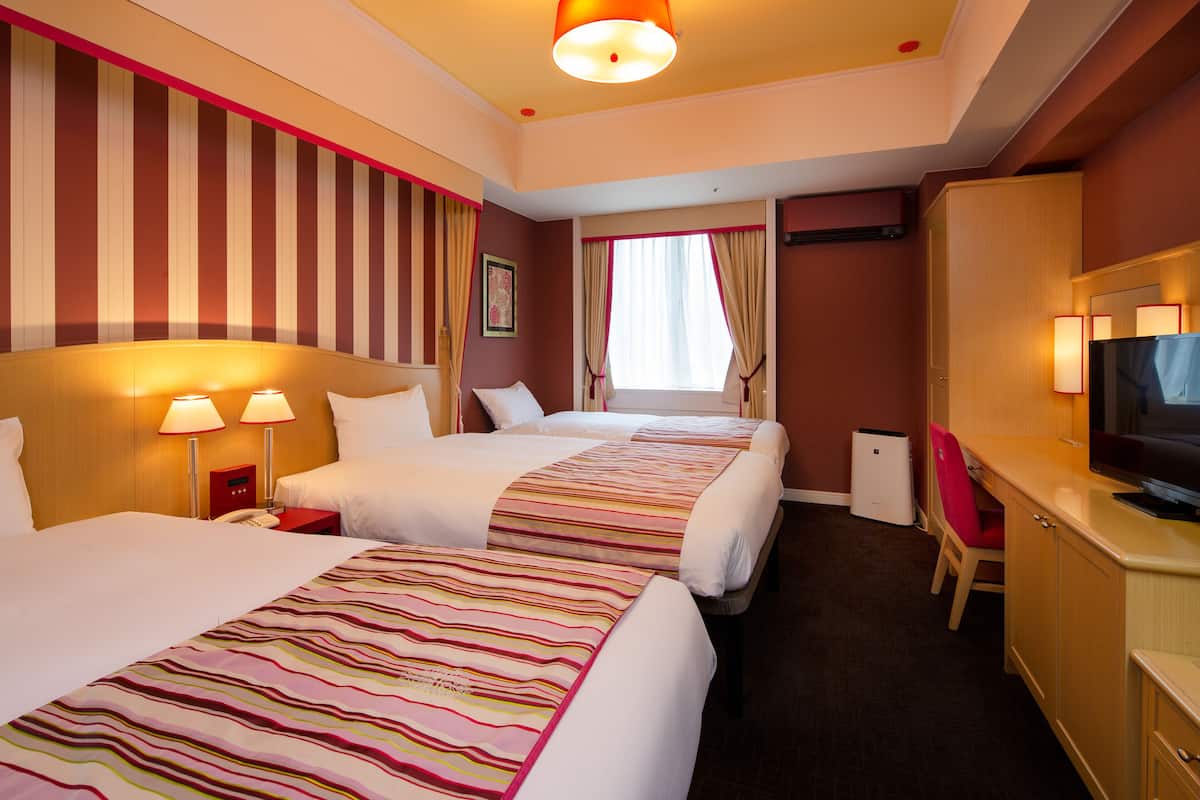 Ein Hotelzimmer mit zwei Betten, einem Schreibtisch, einem Fernseher und einem fensterbepflanzten Fenster.