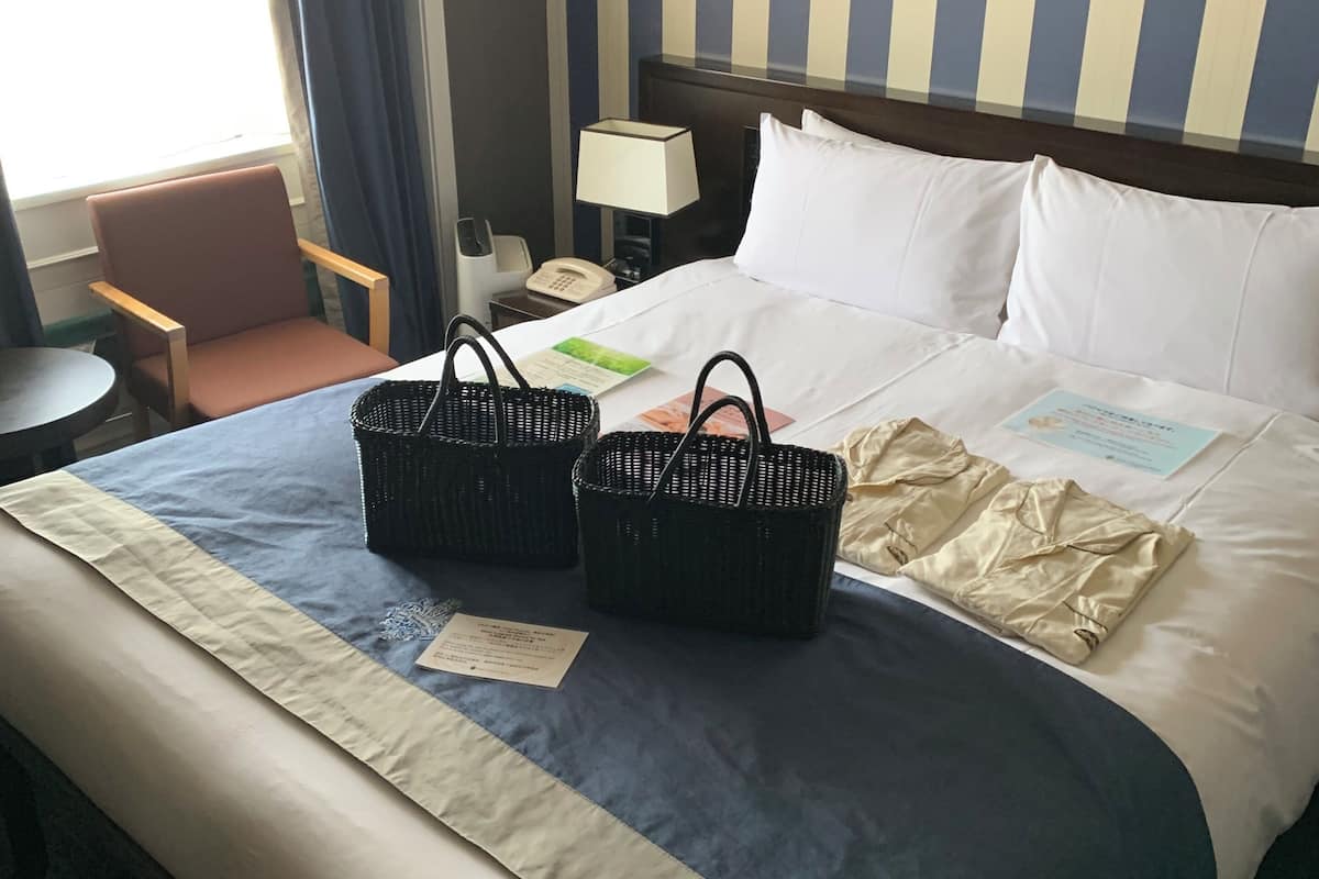 Ein Hotelzimmer mit einem ordentlich bezogenen Bett, zwei schwarzen Körben, einem Nachttisch mit einer Lampe, einem Telefon und einem Fenster mit Vorhängen.