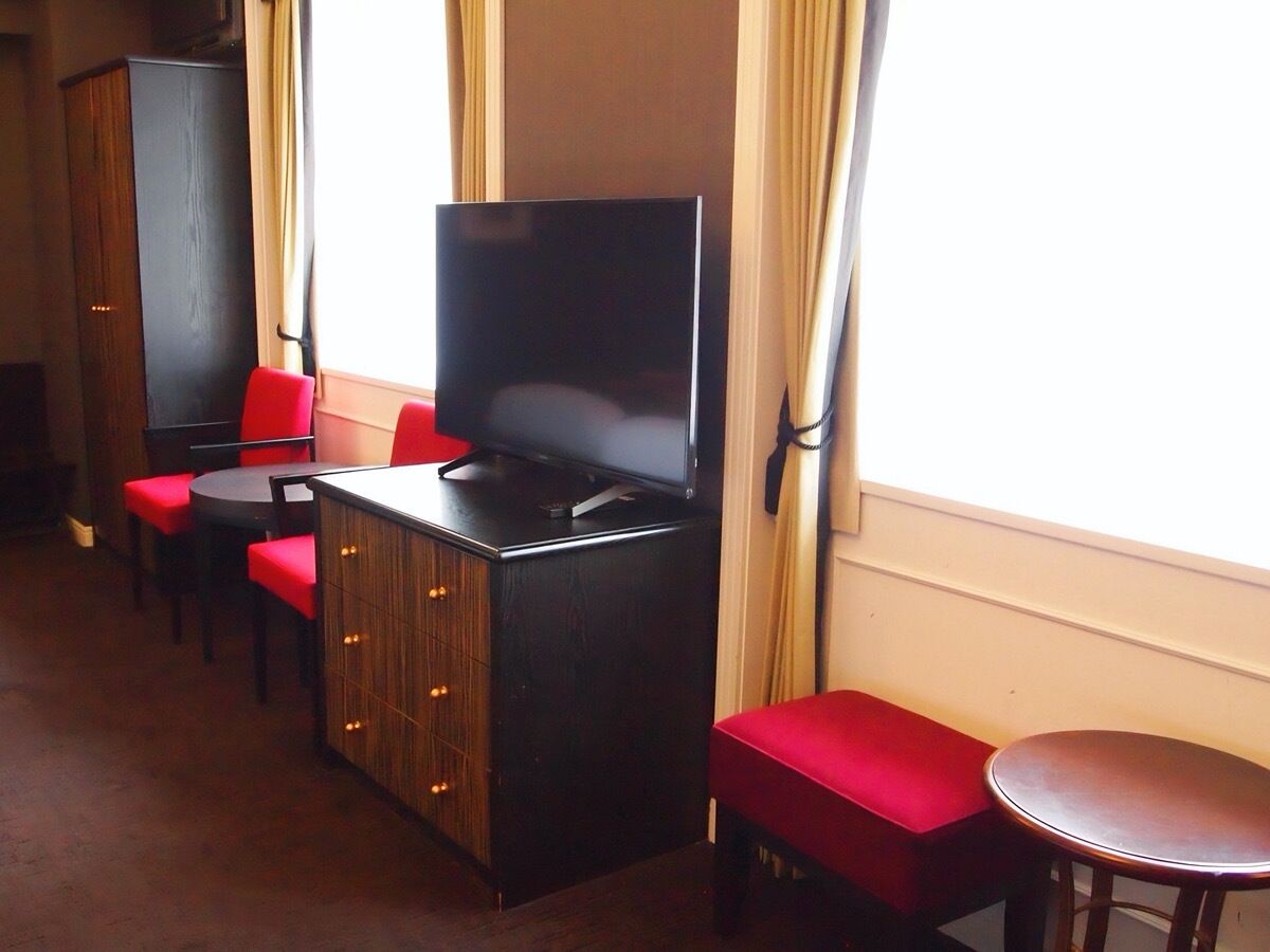 A hotel room with a TV on a wooden cabinet, red seating, and a window with curtains.