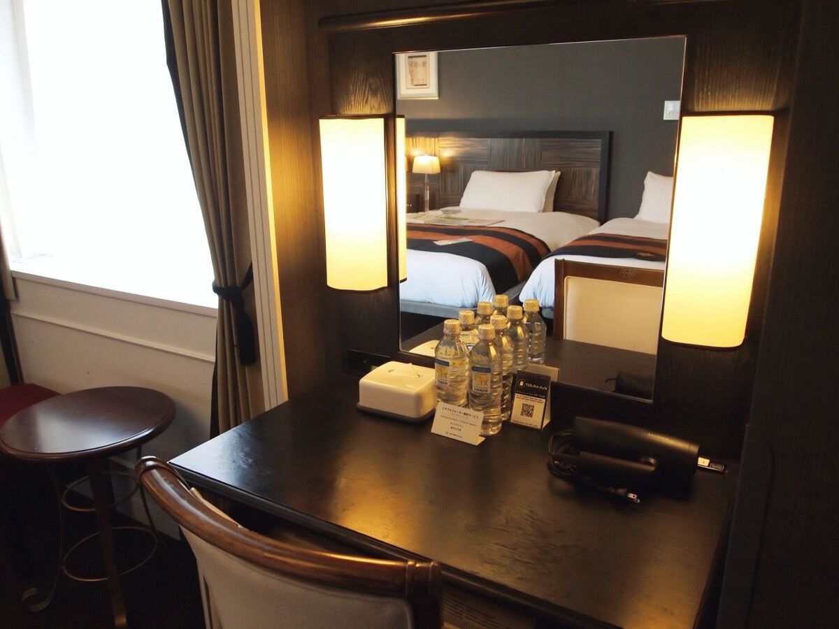 Habitación de hotel con cama, lámparas de noche, un escritorio con teléfono, espejo y ventana con cortinas.