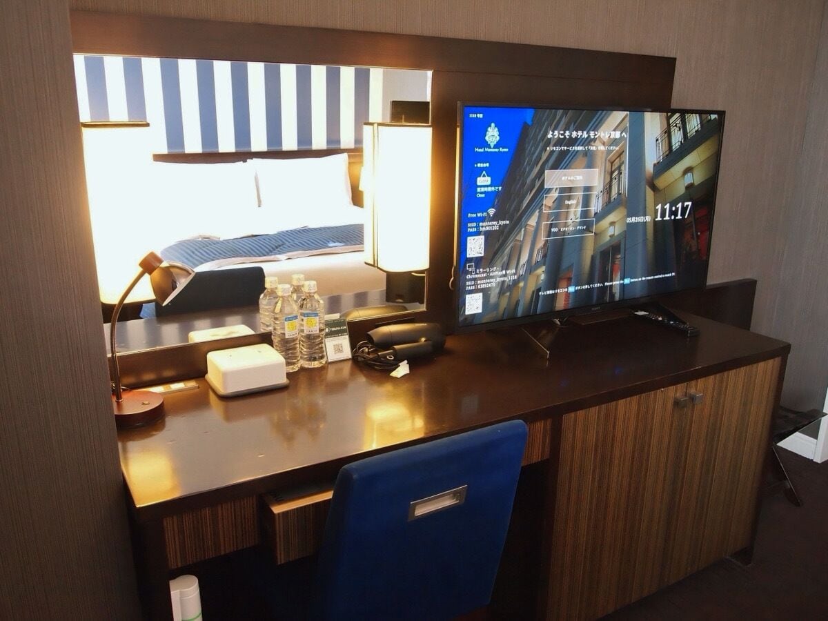 En hotelreception med et fladskærms-tv, der viser et bybillede, en bordlampe, en stol og et lille bord med vandflasker og en servietæller.