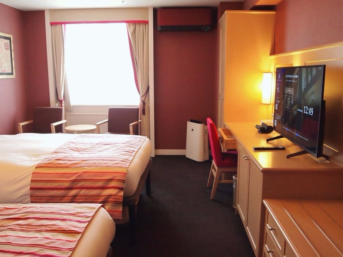 A hotel room with a bed, a desk with a TV, a chair, and a small table.
