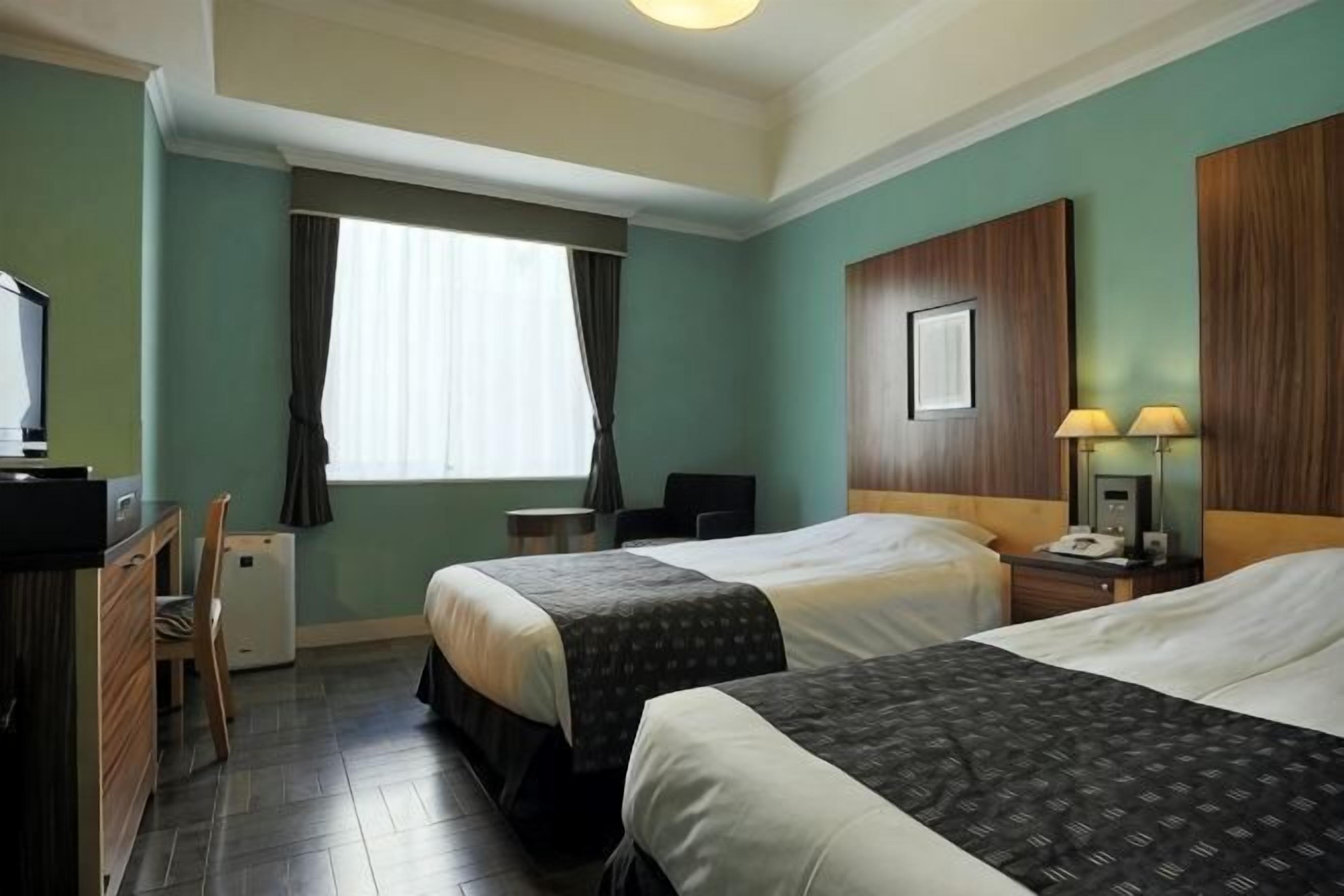 A hotel room with two beds, a desk, a chair, a TV, a window with curtains, and a nightstand with a lamp.