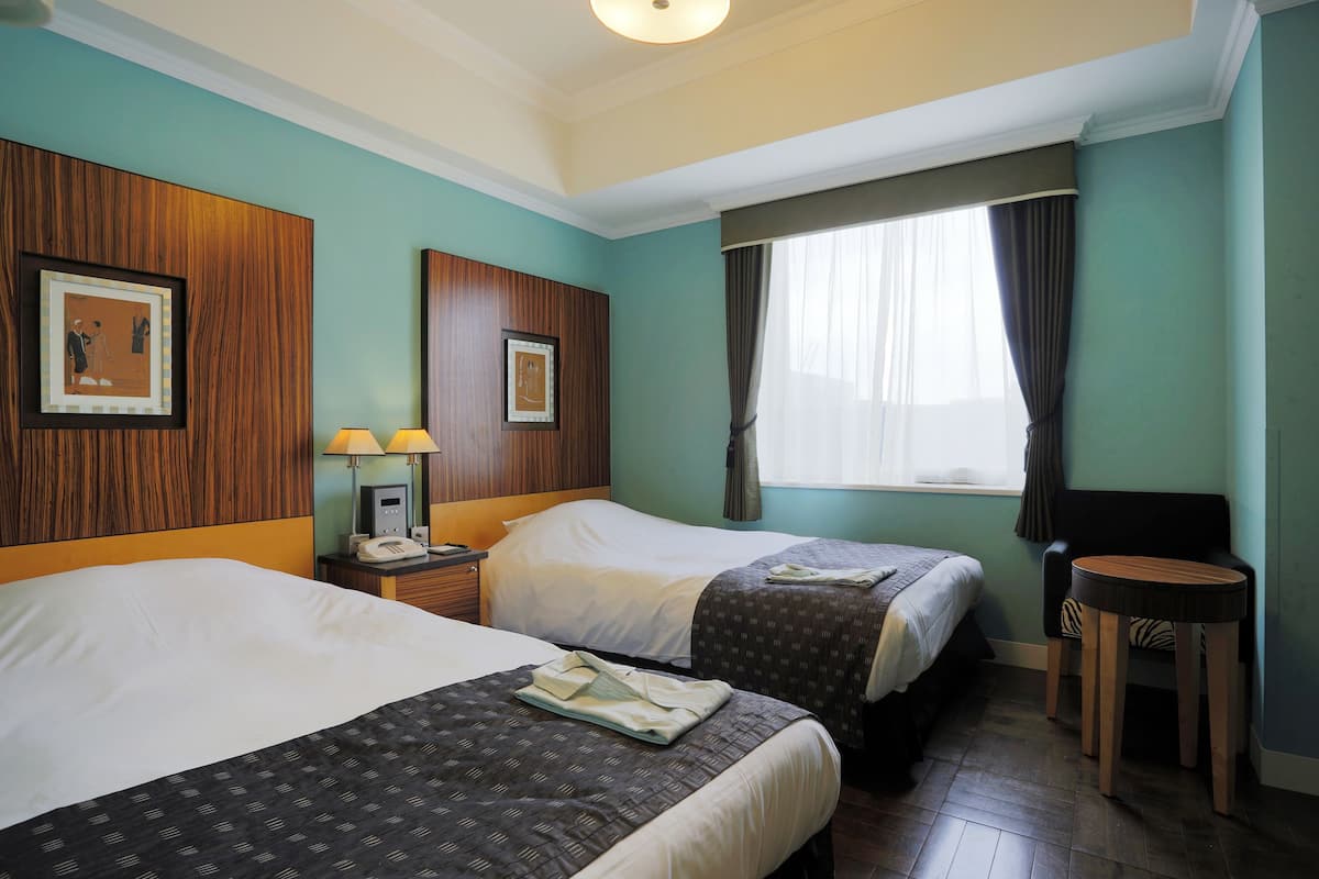 Une chambre d’hôtel avec deux lits, une petite table, une chaise, un bureau avec une lampe et une fenêtre avec des rideaux.