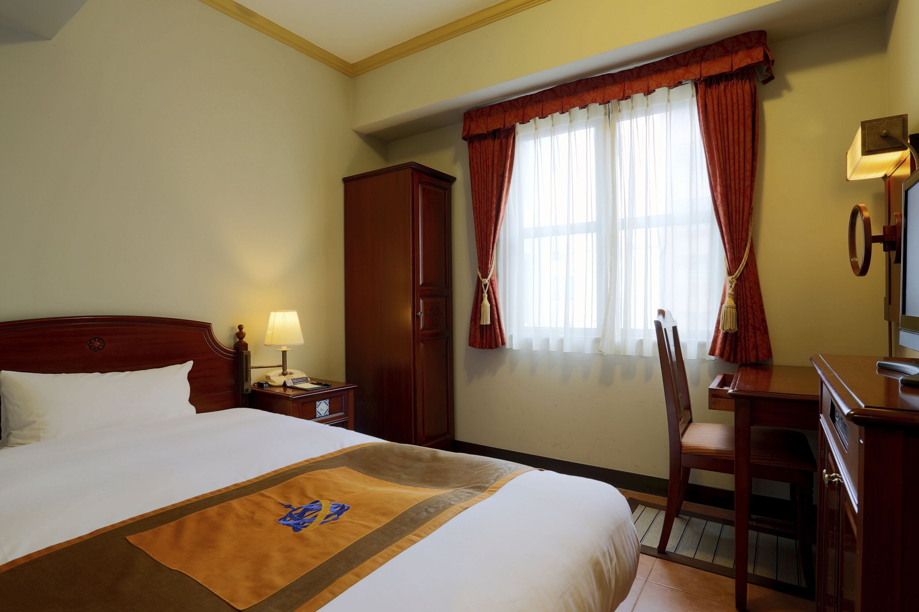 Habitació d'hotel amb un llit gran, una taula de nit de fusta, una cadira, un armari i una finestra amb cortines.