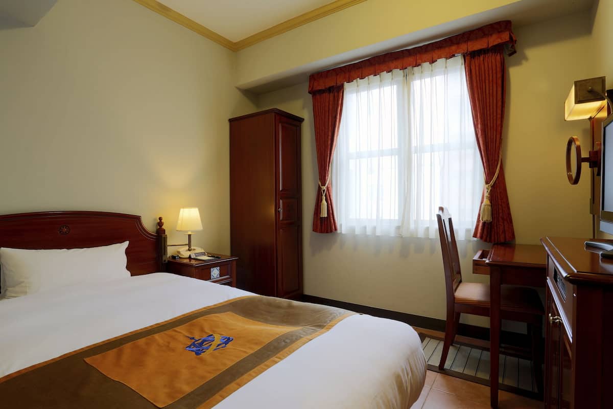 A hotel room with a large bed, a wooden desk, a chair, a wardrobe, and a window with curtains.