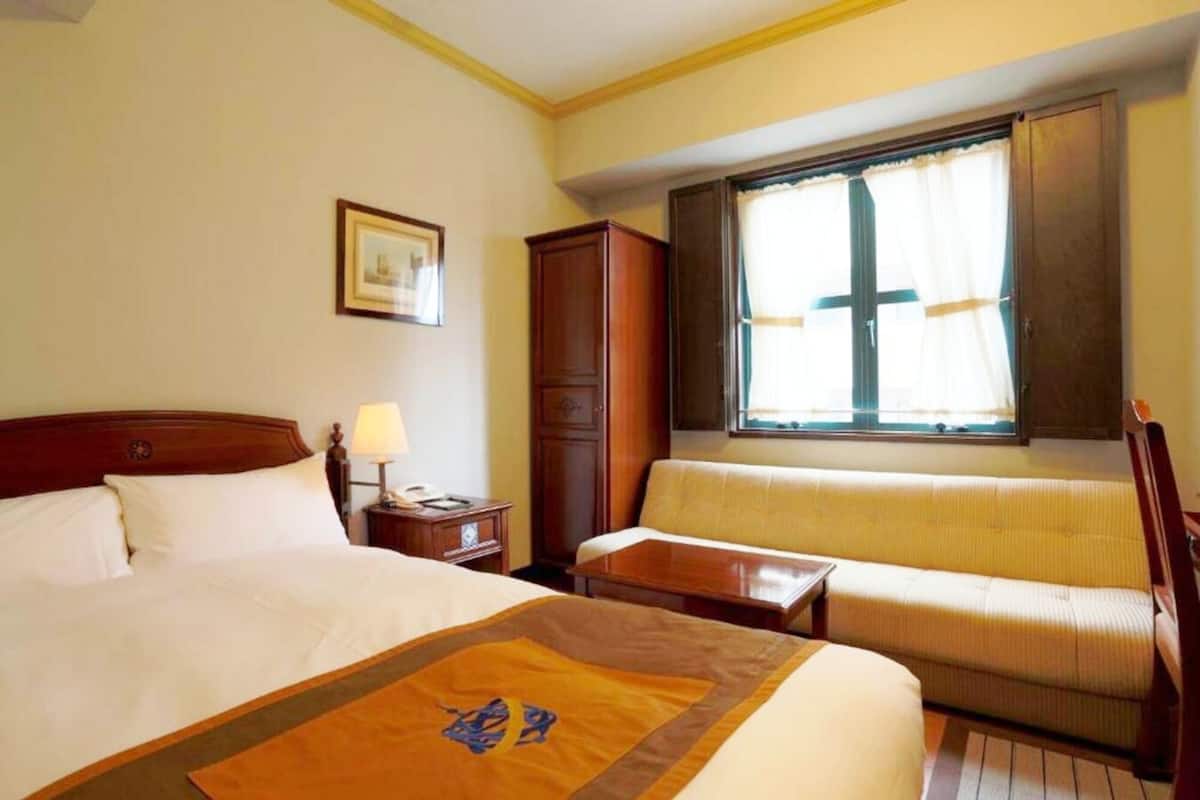 A hotel room with a bed, a sofa, a desk, a lamp, a wardrobe, and a window with curtains.