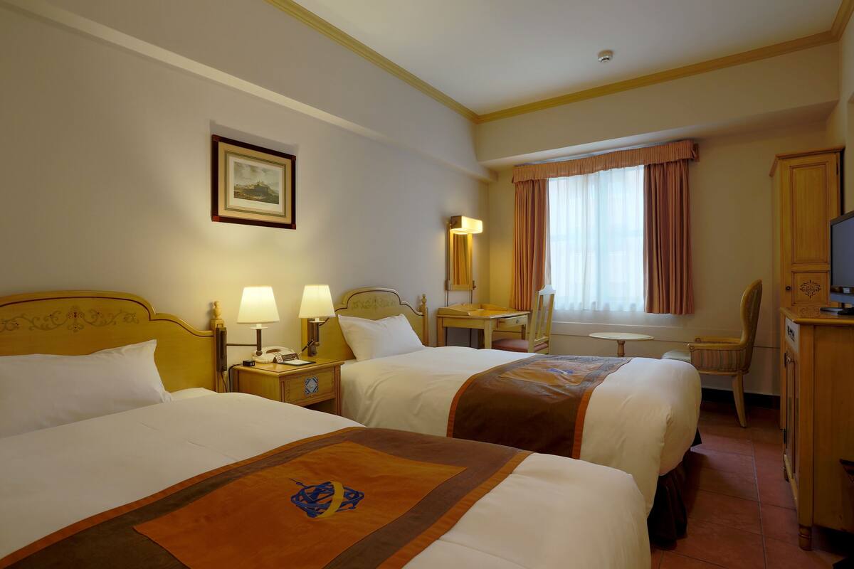Ein Hotelzimmer mit zwei Betten, einem Schreibtisch, einem Stuhl, einem Fernseher und einem Fenster mit Vorhängen.