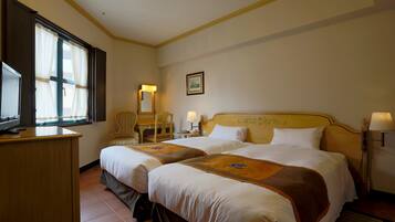 A hotel room with two beds, a desk, a chair, a desk lamp, a bedside table, and a picture on the wall.