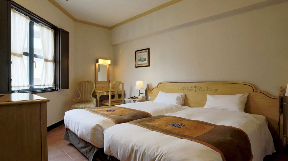 Een hotelkamer met twee bedden, een bureau, een stoel, een nachtkastje met een lamp, en een raam met gordijnen.