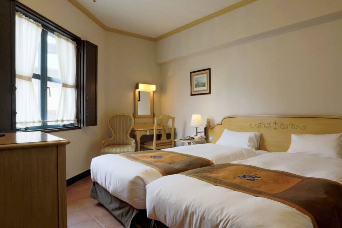 A hotel room with two beds, a desk, a chair, a bedside table with a lamp, and a window with curtains.