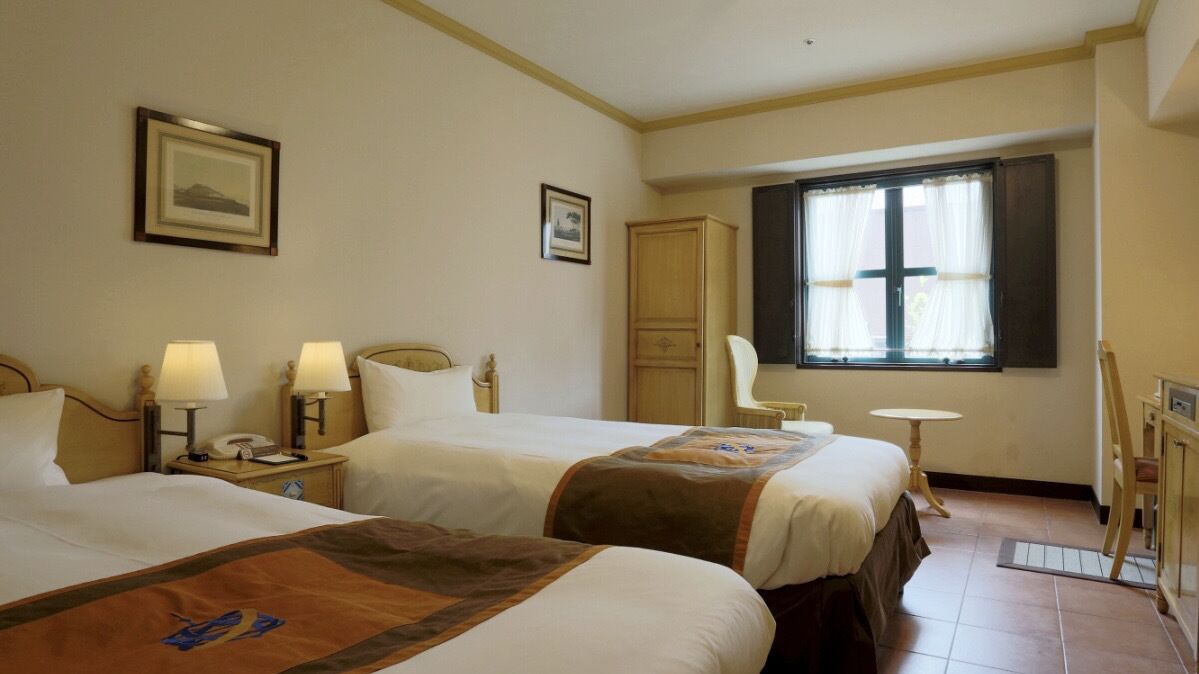 Een hotelkamer met twee bedden, een bureau, een stoel, een kledingkast en een raam met gordijnen.