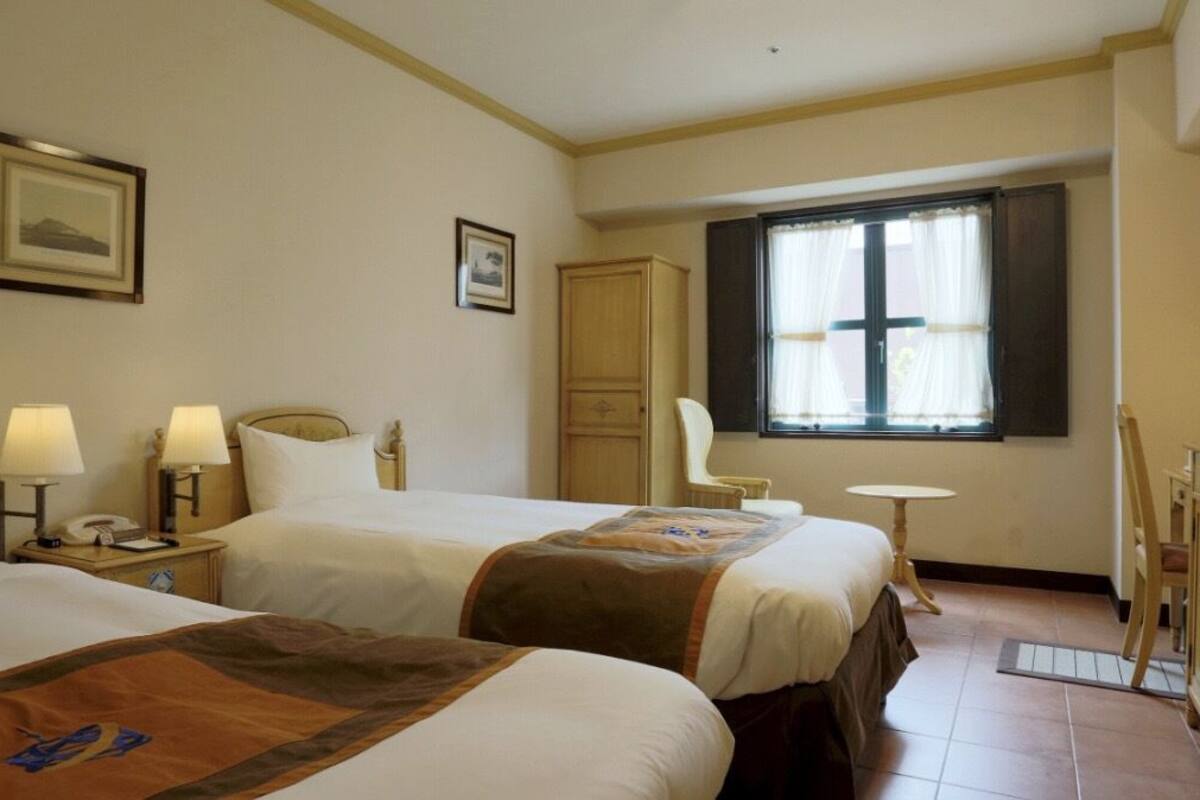 Ein Hotelzimmer mit zwei Betten, einem Schreibtisch, einem Stuhl, einem Kleiderschrank und einem Vorhangfenster.