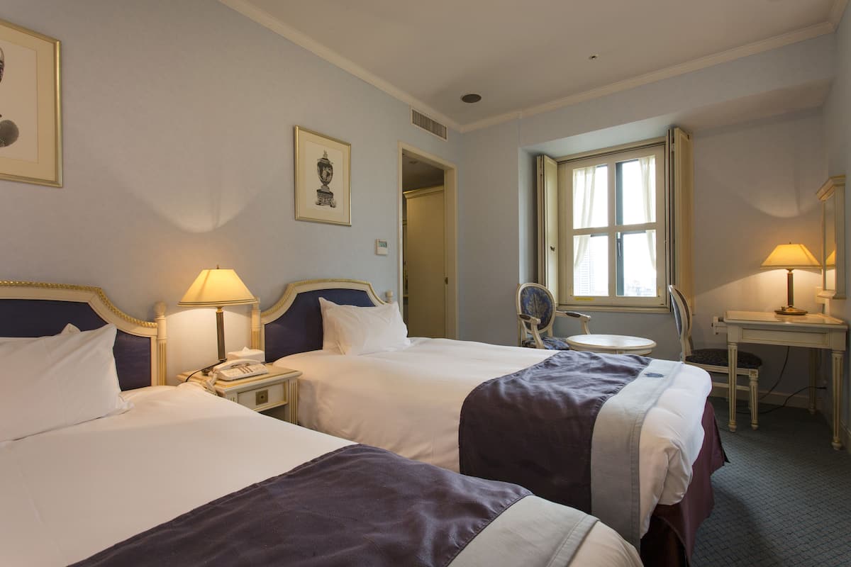 A hotel room with two beds, a small table, a chair, and a window with curtains.