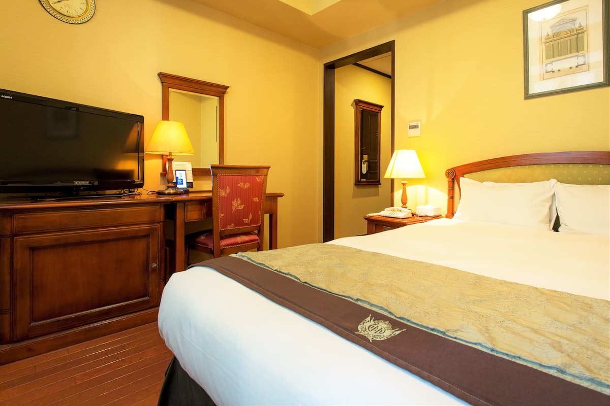A hotel room with a bed, a desk with a television, a chair, a nightstand with a lamp, and a clock on the wall.