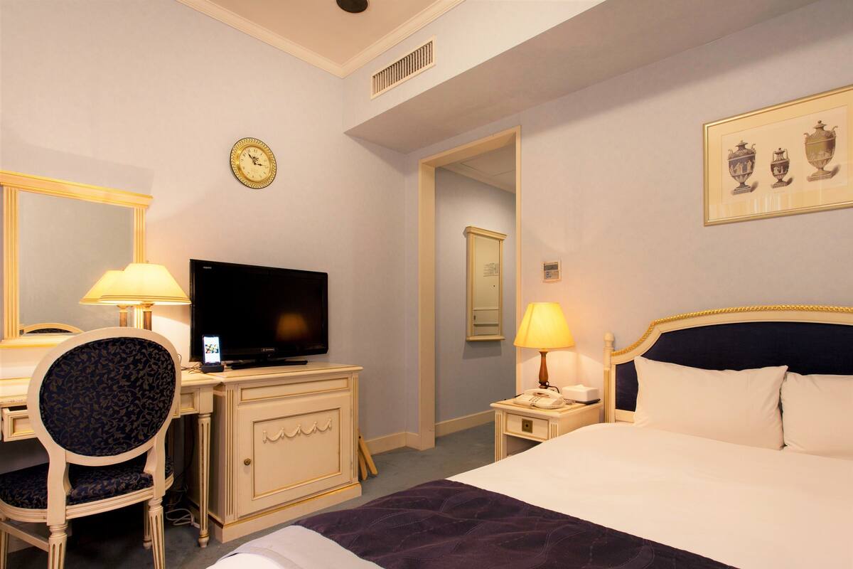 A hotel room with a bed, a desk with a computer, a chair, a television, a clock, and a framed picture on the wall.