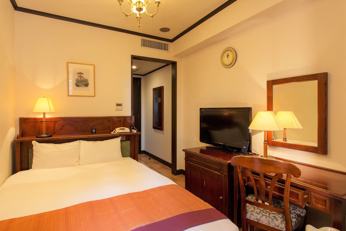 A hotel room with a bed, bedside table, TV, desk, chair, and a mirror.