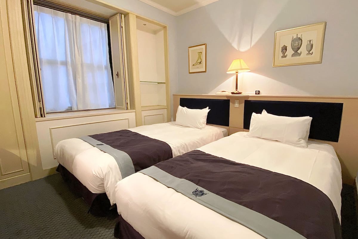 A hotel room with two beds, a window with curtains, a bedside table with a lamp, and two framed pictures on the wall.