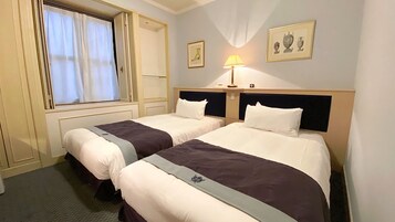 Quarto de hotel com duas camas, uma janela com cortinas, uma mesa de cabeceira com abajur e duas imagens emolduradas na parede.