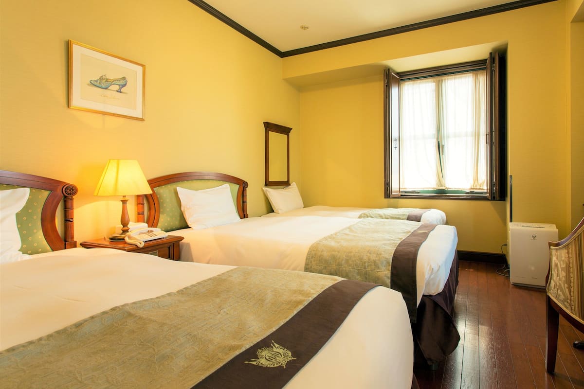A hotel room with two beds, a bedside table with a lamp, a framed picture on the wall, and a window with curtains.
