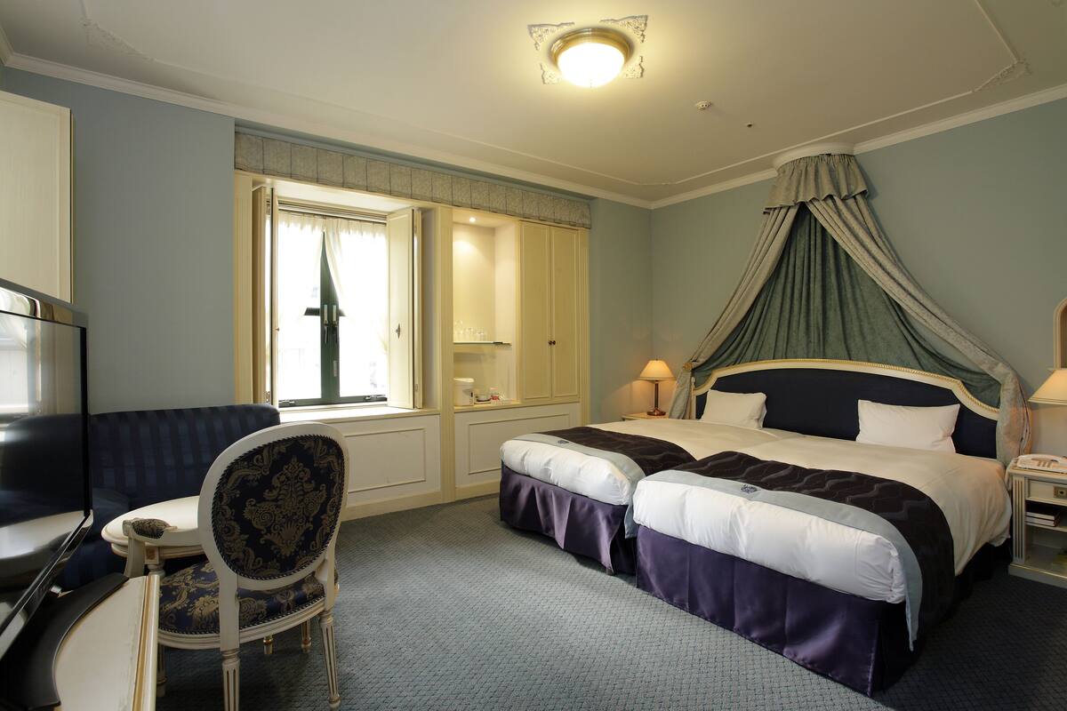 A hotel room with two beds, a desk, a chair, a TV, and a window with curtains.