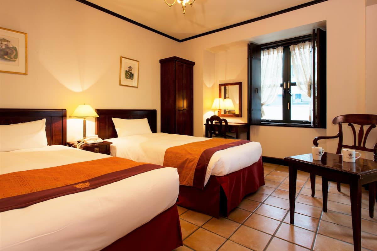 A hotel room with two beds, a desk, a chair, a lamp, and a window with curtains.