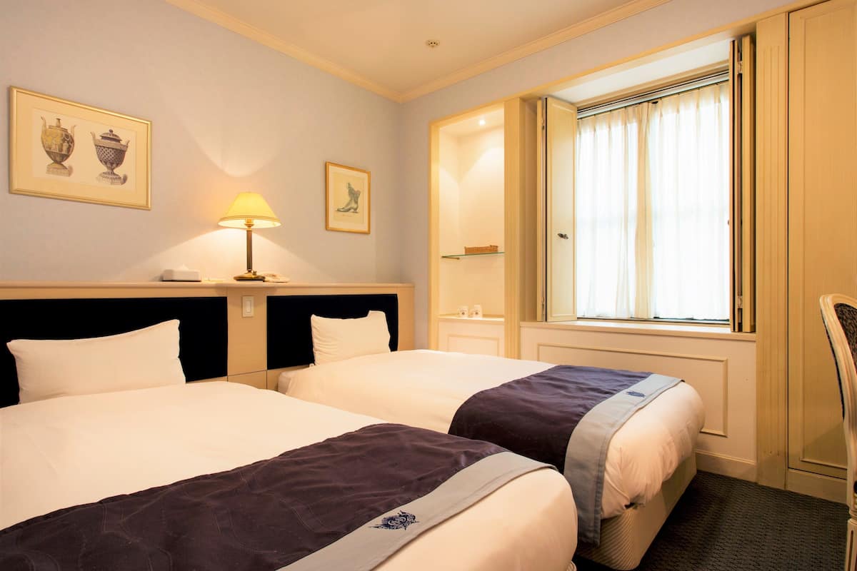 A hotel room with two beds, a bedside table with a lamp, a framed picture on the wall, and a window with curtains.