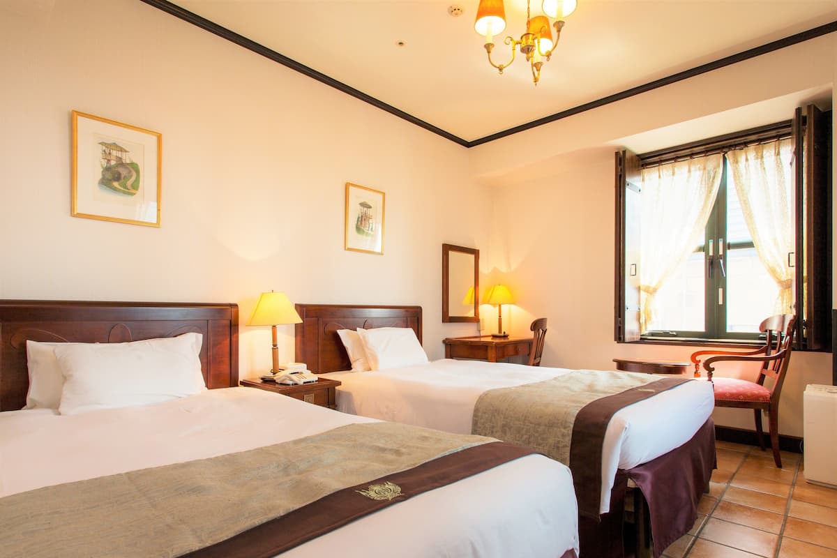 A hotel room with two beds, a desk, a chair, a window with curtains, and a chandelier.