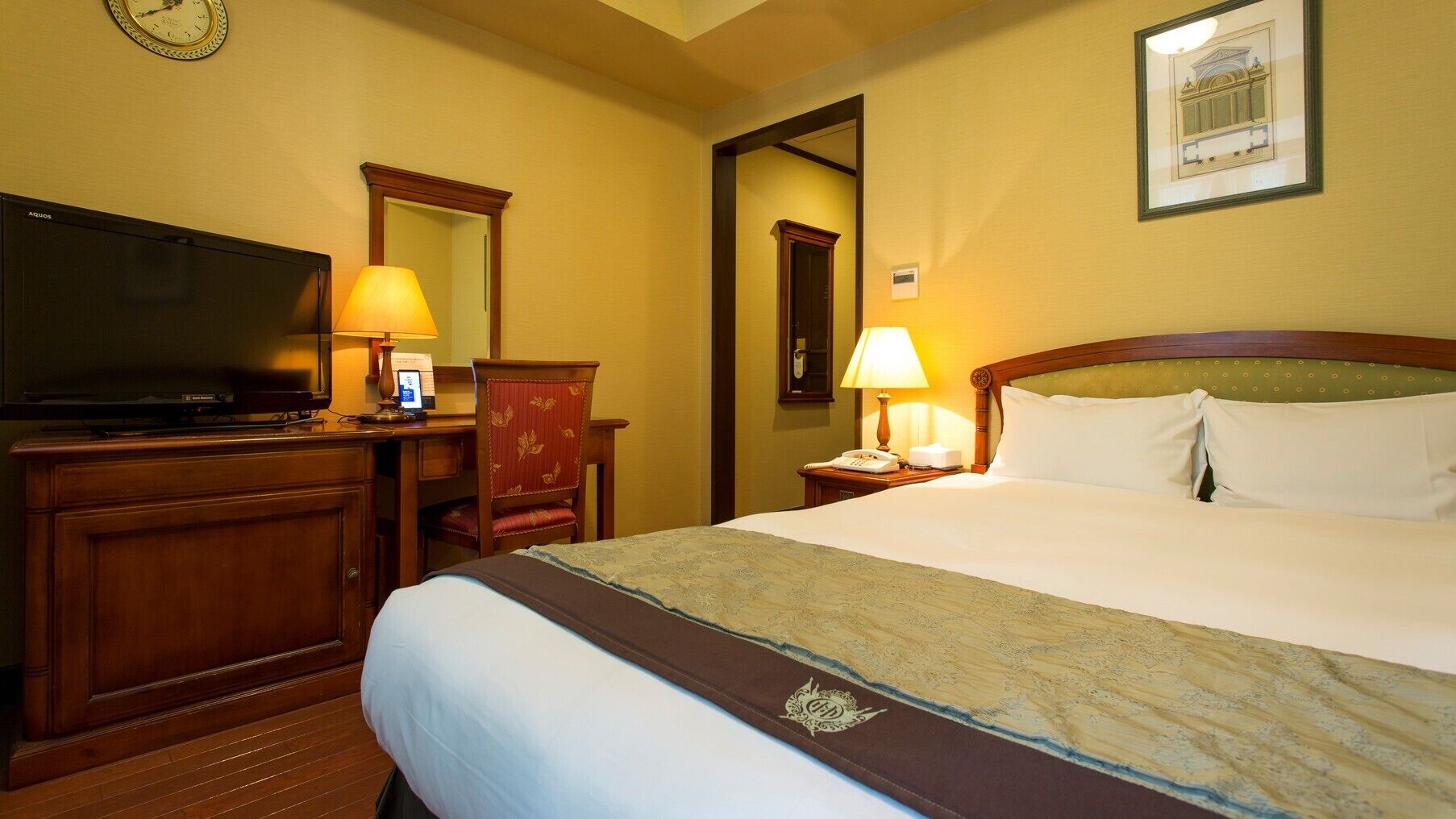 A hotel room with a bed, a desk with a television, a chair, a nightstand with a lamp, and a framed picture on the wall.