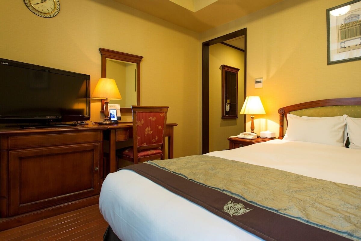 A hotel room with a bed, a desk with a television, a chair, a nightstand with a lamp, and a framed picture on the wall.