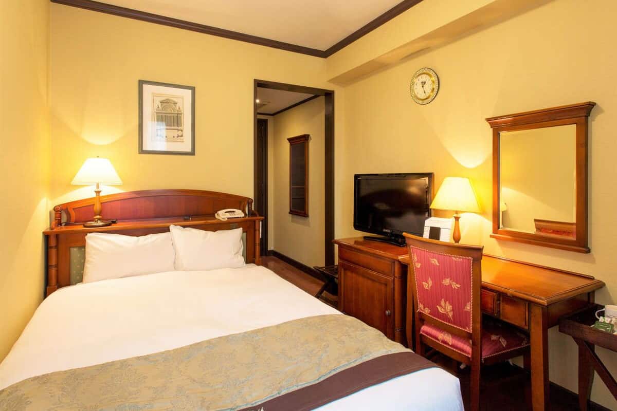 A hotel room with a large bed, bedside table, lamp, TV, desk, chair, and a mirror.