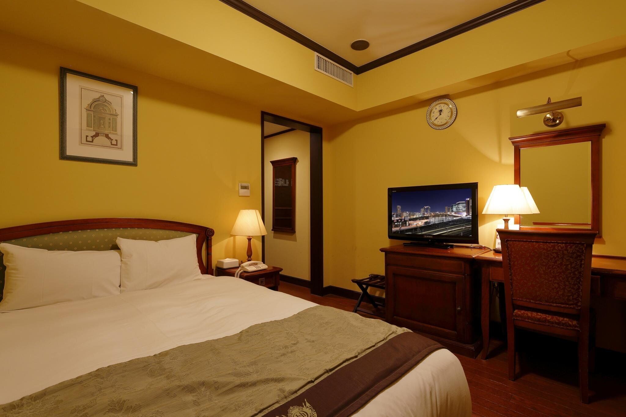 Ein Hotelzimmer mit Bett, Fernseher, Schreibtisch mit Lampe, Sessel und einer Wanduhr.