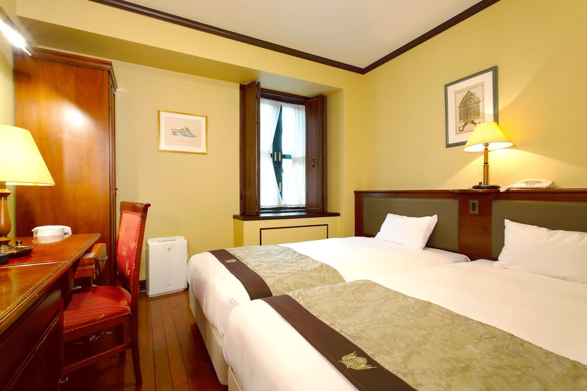 A hotel room with two beds, a desk with a lamp, a chair, a window with curtains, and a framed picture on the wall.