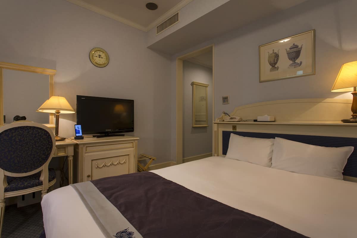 A hotel room with a bed, a desk with a television, a chair, a clock, and a framed picture on the wall.