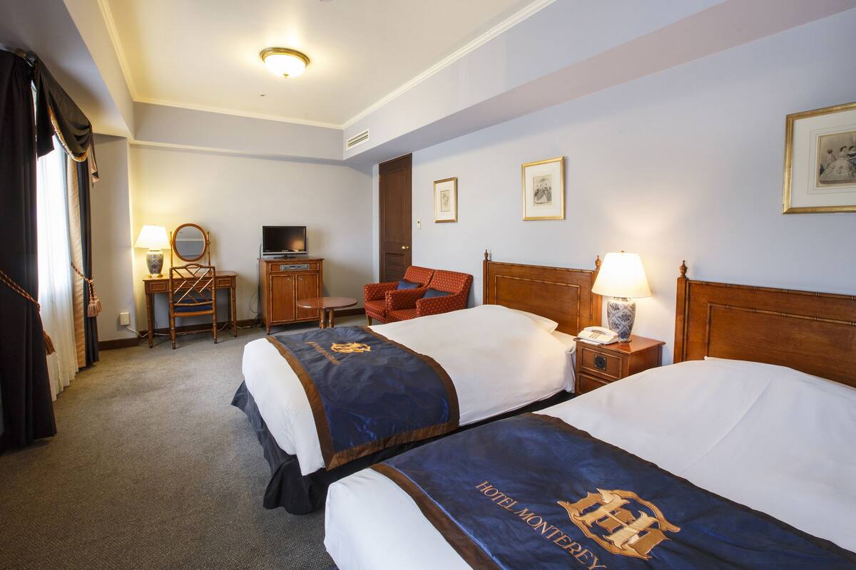 A hotel room with two beds, a desk, a chair, a television, and framed pictures on the wall.