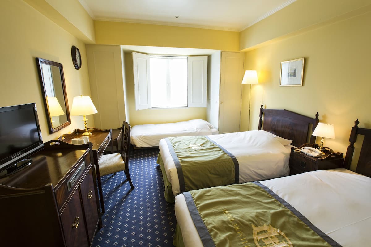 A hotel room with two beds, a TV, a desk with a lamp, and a window.