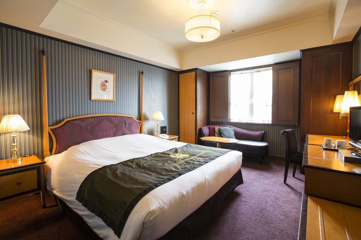 A hotel room with a large bed, a desk, a chair, a sofa, a window, and a lamp.