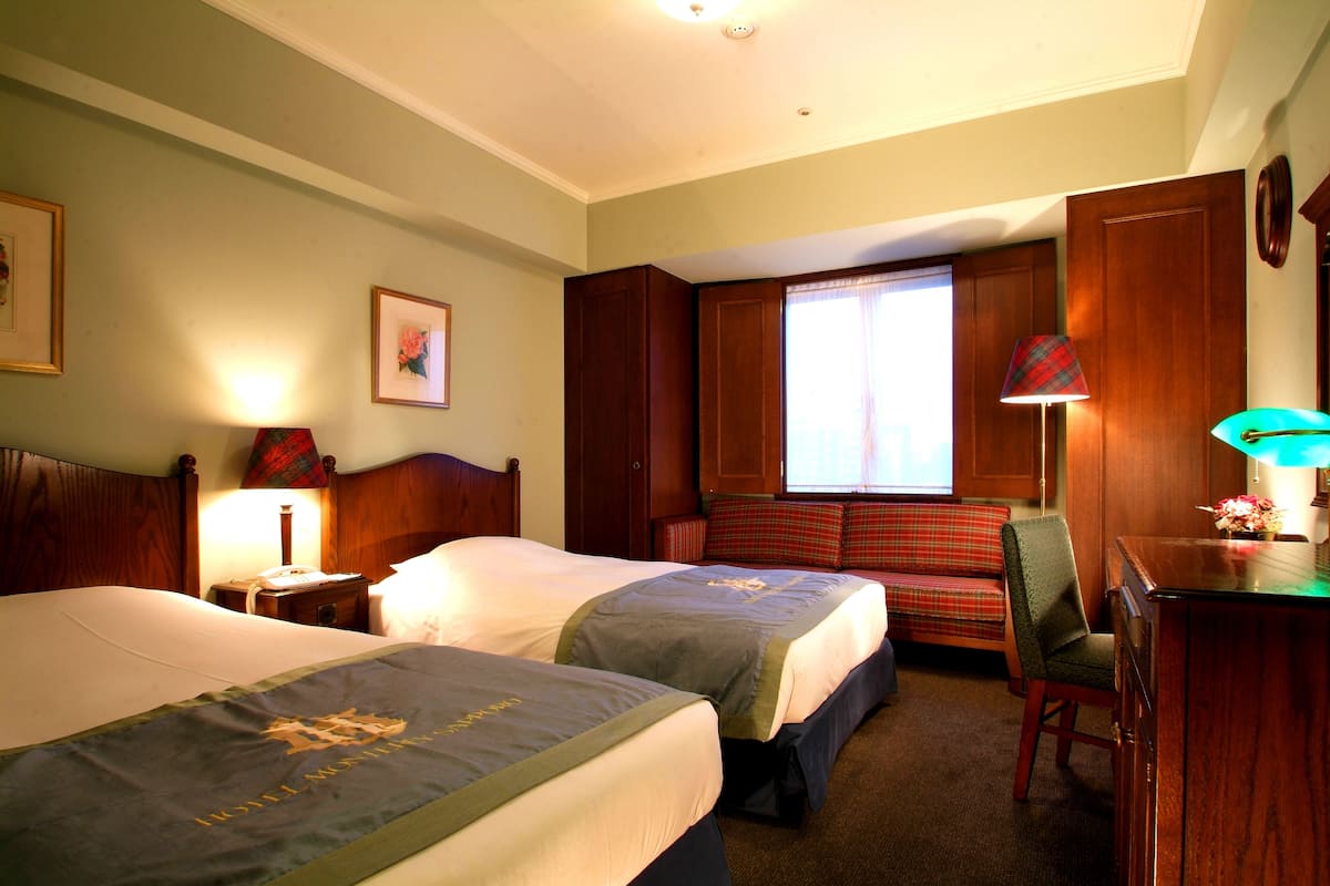 A hotel room with two beds, a desk, a chair, a window with curtains, and a lamp.