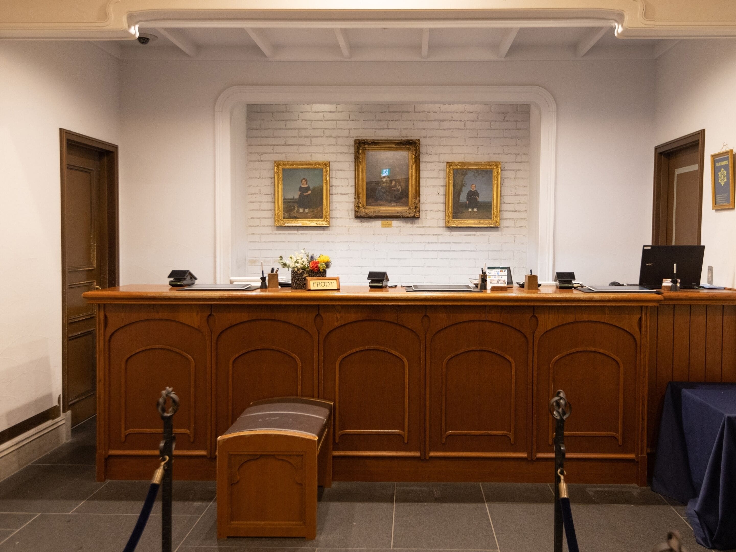 A reception desk with a white brick wall behind it, framed paintings, a wooden counter, and a small bench.