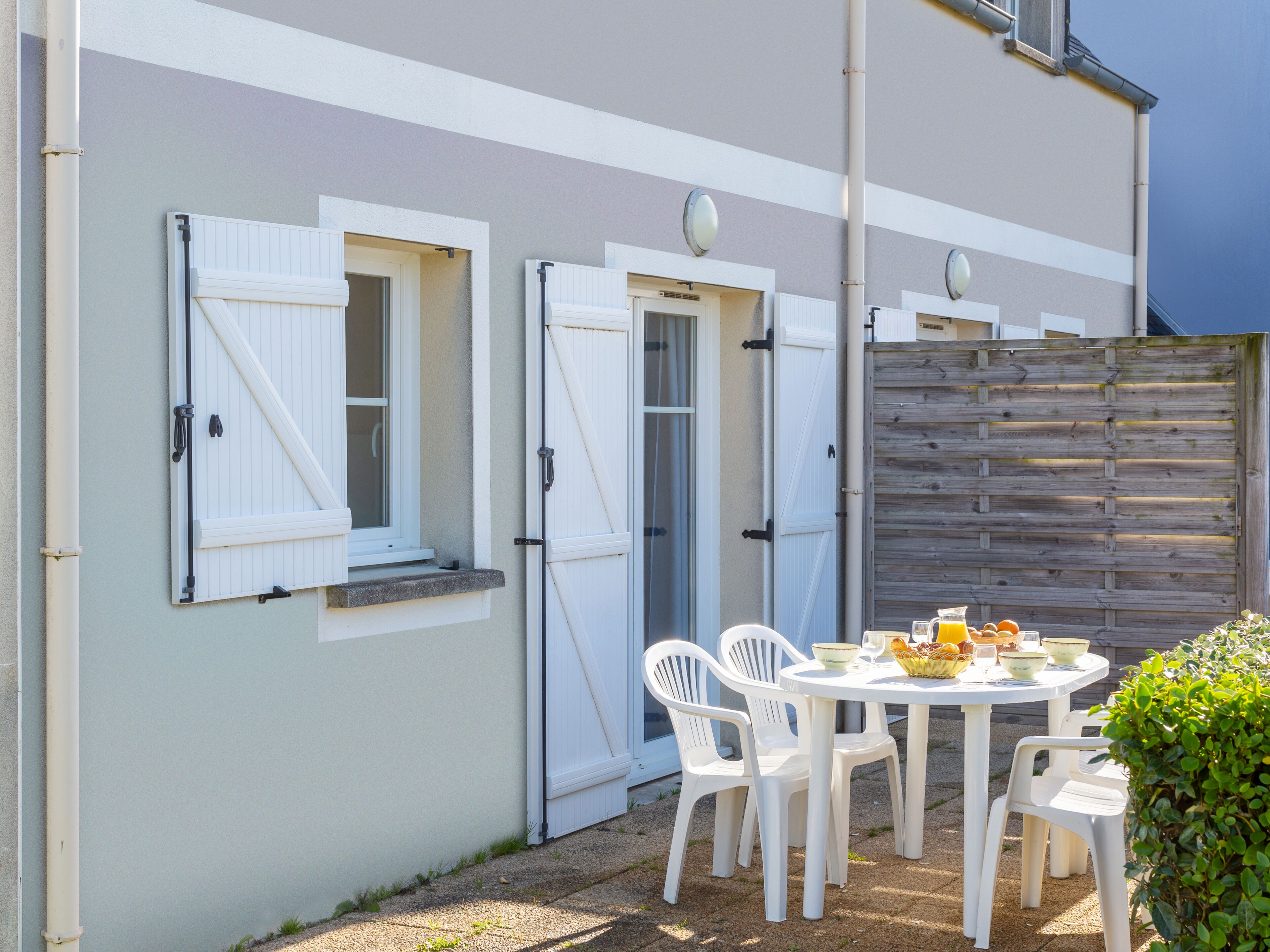 A patio with a round table and white plastic chairs, a wooden fence, and a white building with shutters.