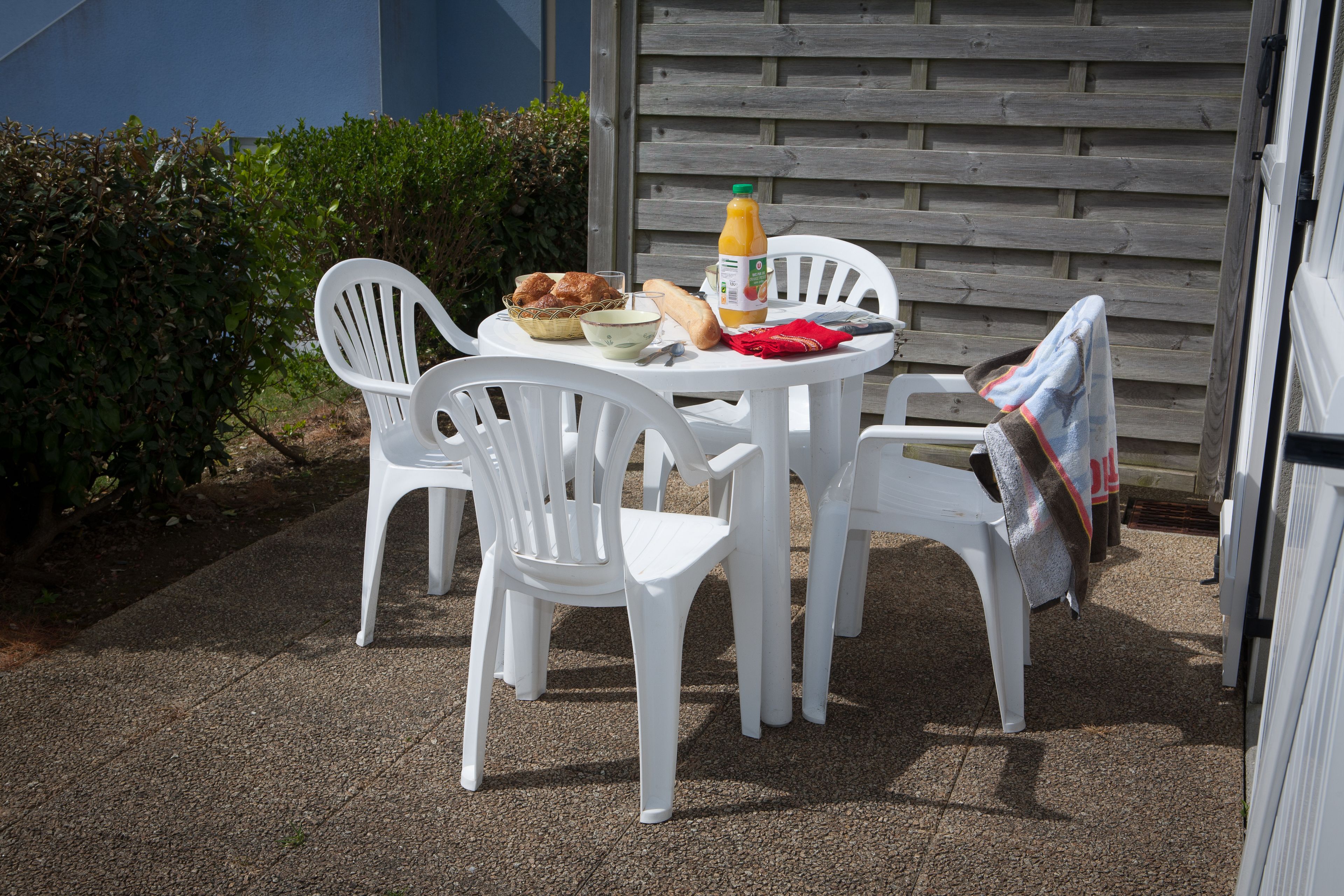 Un patio con sedie e tavolo in plastica bianca, un cesto di pane, una ciotola, una bottiglia di succo d'arancia e un panno rosso.