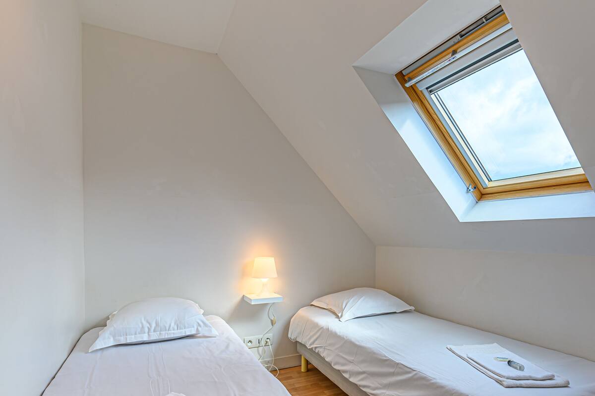 Una habitación pequeña y luminosa con dos camas, un tragaluz y una lámpara de noche.