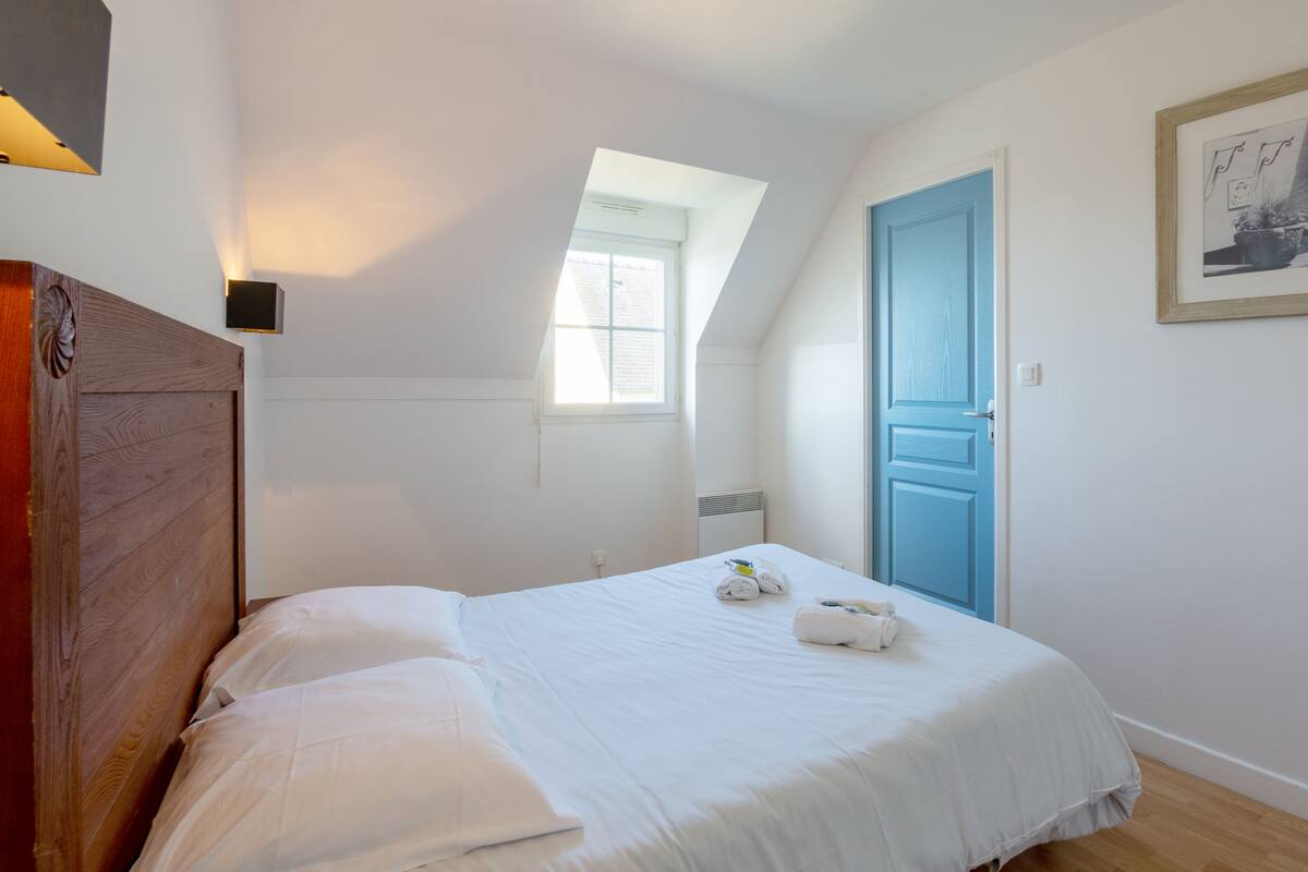 Un dormitorio con techo inclinado, tragaluz, puerta azul y cama con ropa de cama blanca.