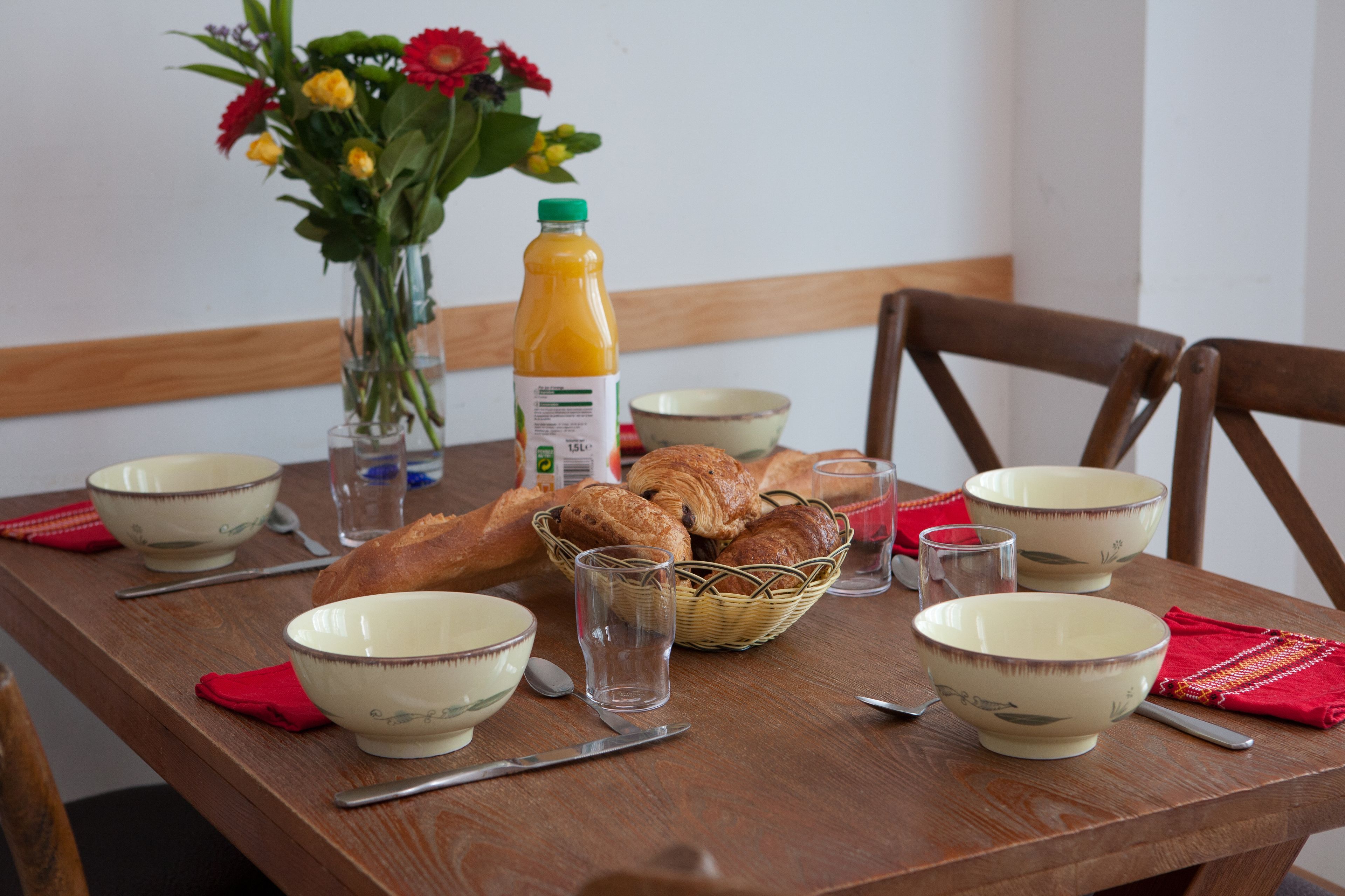 A wooden dining table with bowls, a glass of juice, a basket of bread, and a vase with flowers.