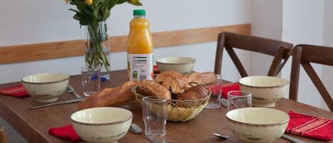 A wooden dining table with bowls, a glass of juice, a basket of bread, and a vase with flowers.